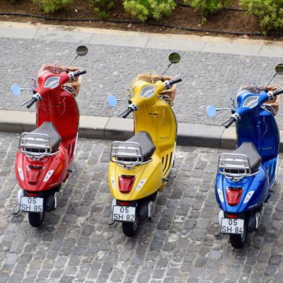 Adhetec regelt met slimme camera's ook kentekenplaatherkenning. Sfeerbeeld van een rode, gele en blauwe eenpersoons scooter die langs de weg zijn geparkeerd. Ze hebben alledrie een rietenmandje voorop en staan met het stuur naar links gedraaid.