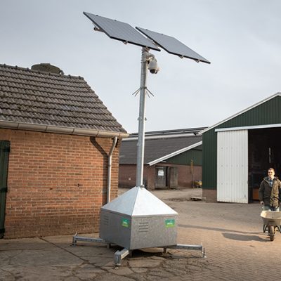 Adhetec camerabewaking op het erf Sfeerbeeld van een boerenerf met drie schuren en voor de eerste staat een mobiele cameramast. De boer komt met een kruiwagen aanlopen.