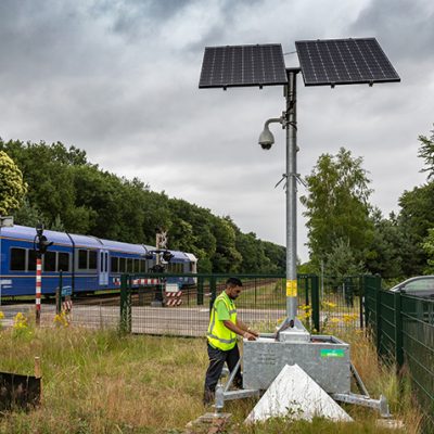 Sfeerbeeld van spoorwegveiligheid, want op de groenstrook langs het spoor staat bij een onbewaakte spoorwegovergang een mobiele cameramast voor toezicht. Er naast staat een man met een geel veiligheidsvestje onderhoudswerkzaamheden uit te voeren. Er komt ook een blauwe trein langsrijden en er staat een zwarte personenauto voor de slagbomen te wachten.