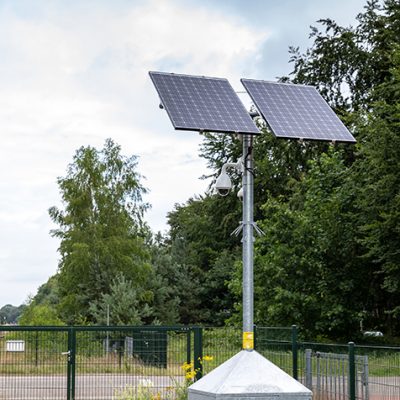 Sfeerbeeld van camerabeveiliging met een mobiele cameramast die is opgebouwd uit een ijzeren bak met accu's daarboven hangen in een ijzeren paal beveiligingscamera's met daarboven twee zonnepanelen.