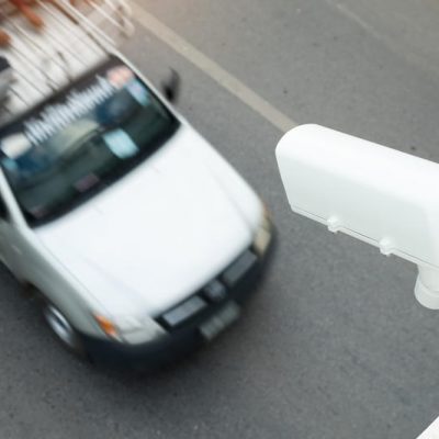 Sfeerbeeld van bovenaf een wegovergang genomen. Er rijdt een witte auto onder een verkeerscamera door.