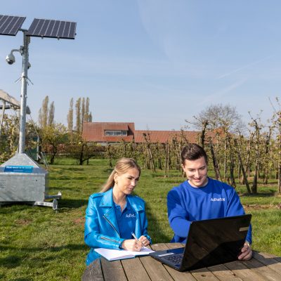Mobiele cameramast van Adhetec in een boomgaard Sfeerbeeld van camerabeveiliging met een mobiele cameramast met zonnepanelen in een boomgaard. Op de voorgrond zitten een man en een vrouw aan een picknicktafel en kijken naar een laptop.